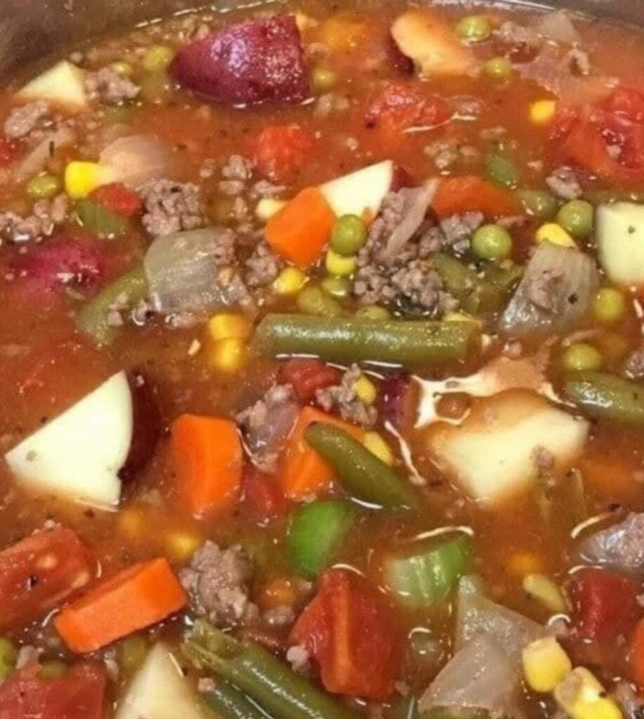 A steaming bowl of hearty ground beef vegetable soup with carrots, potatoes, corn, peas, and green beans in a rich tomato broth.