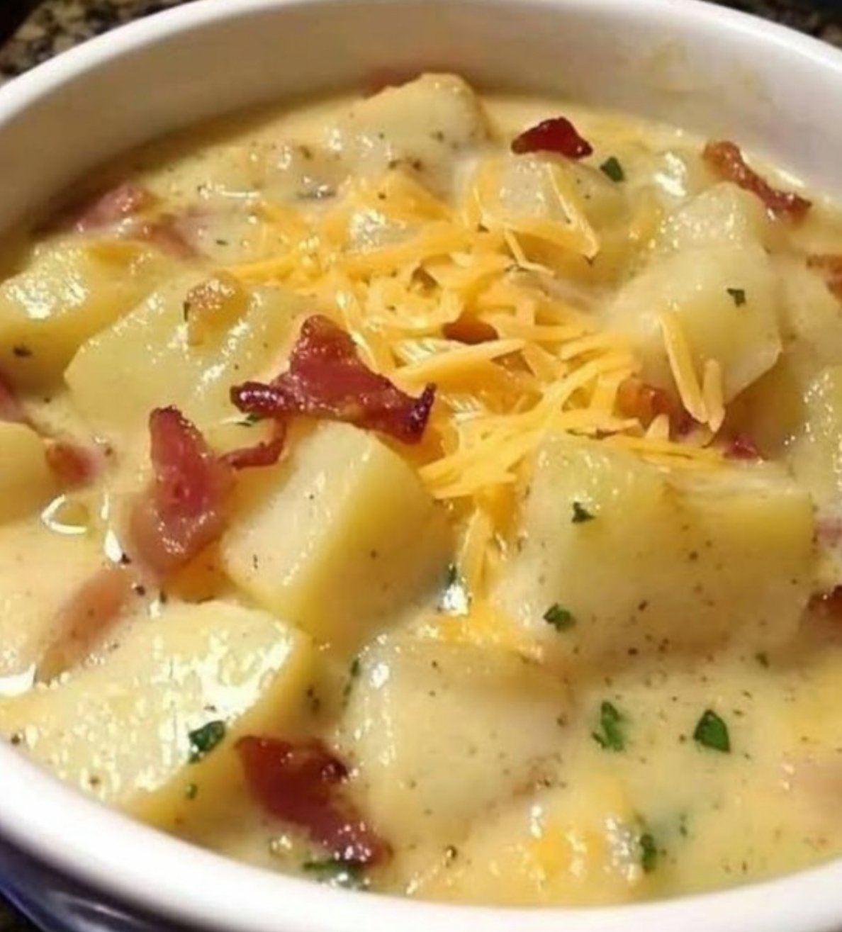A bowl of creamy loaded potato soup topped with crispy bacon, shredded cheddar cheese, and sliced green onions, served with crusty bread on the side.